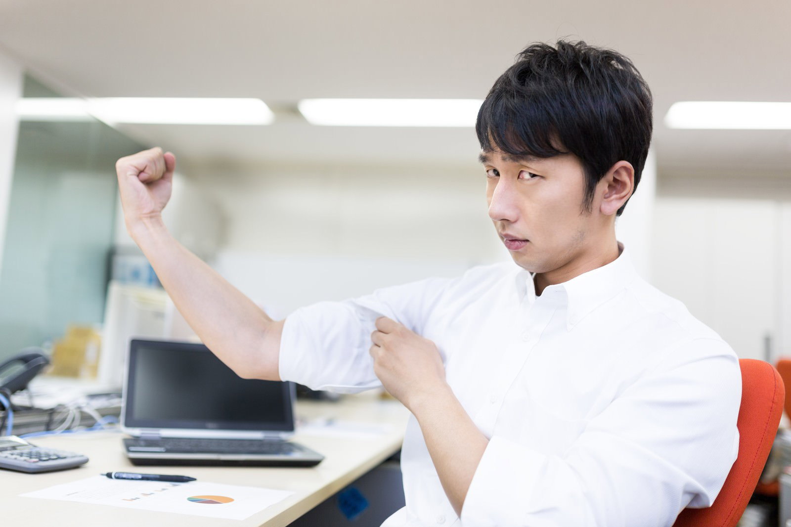 白いTシャツの男性がオフィスデスクで左腕を上げ力こぶをアピール［モデル：大川竜弥］