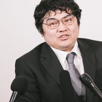 マイクを前に話す記者会見の広報男性、スーツ姿で企業発表の写真