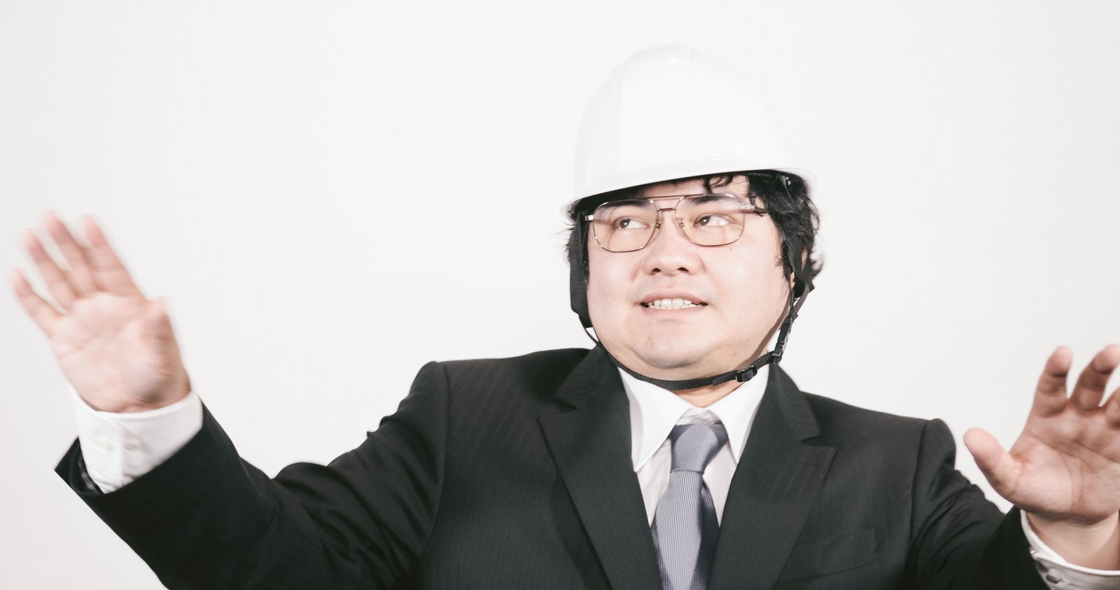 白いヘルメットをかぶったスーツ姿の男性職員が両手を上げて冷静を呼びかけているポーズ［モデル：段田隼人］