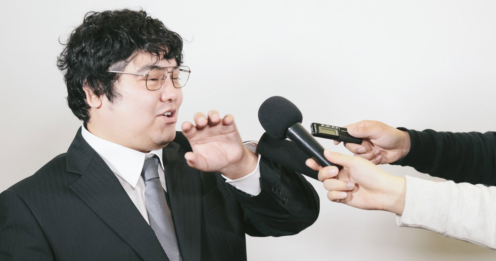 複数のマイクを向けられるスーツ姿の会社役員［モデル：段田隼人］