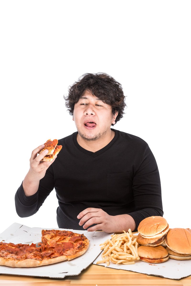 テーブルの上のピザやハンバーガーを前に、疲れた表情で口を開けている男性［モデル：朽木誠一郎］