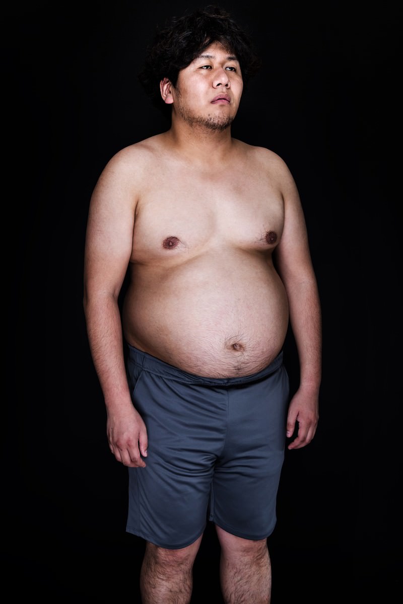 上半身を露出した太めの男性が黒背景で正面を向いて立つ写真［モデル：朽木誠一郎］