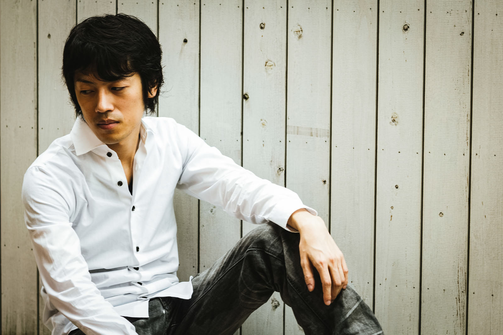 木目調の壁を背景に床に座り白いシャツを着た男性［モデル：Tsuyoshi.］