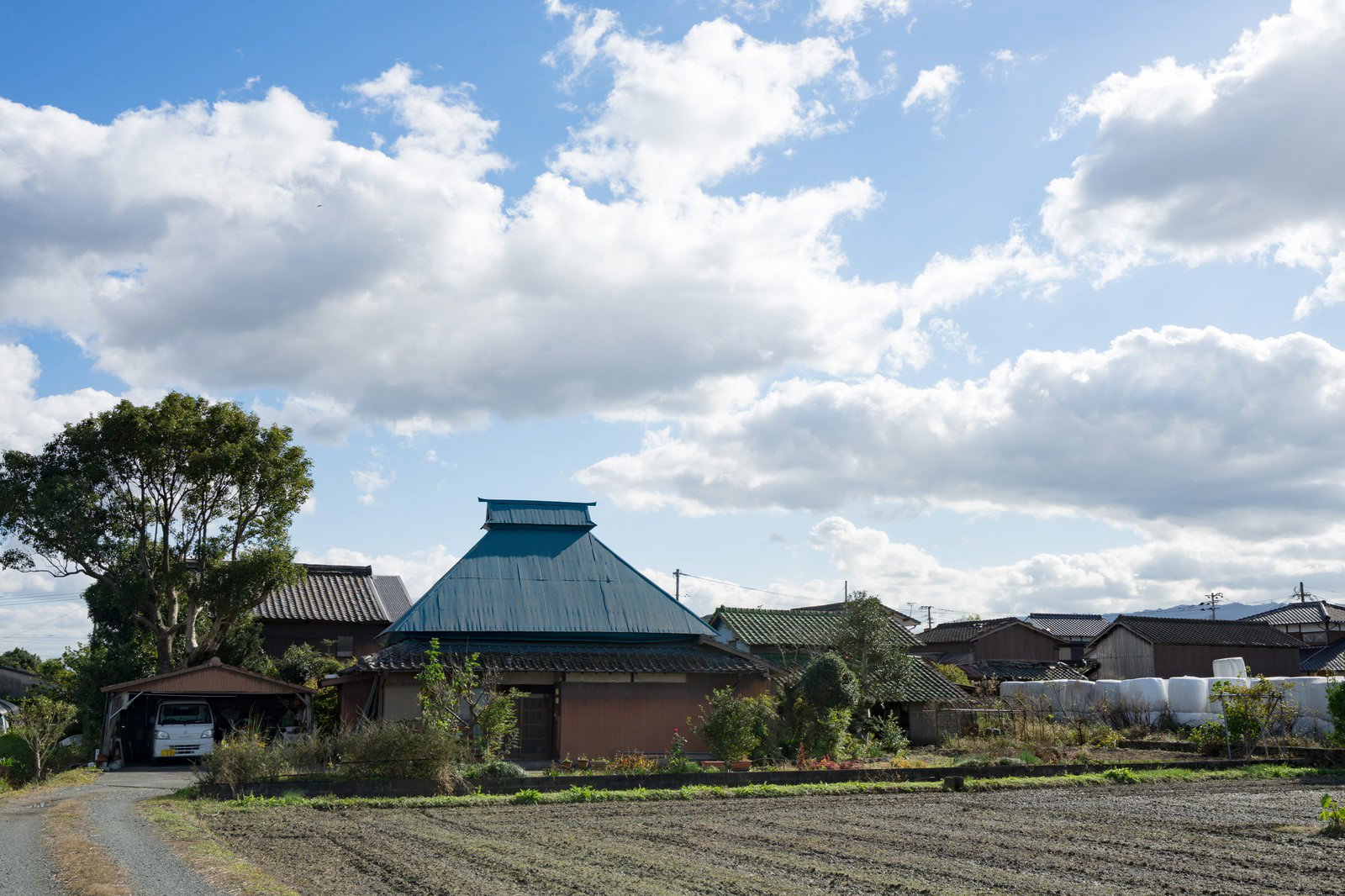 青い屋根の古民家と瓦葺き建屋が立つ敷地。前方に耕作地が広がり、左側に大きな樹木がある福岡県の農村風景