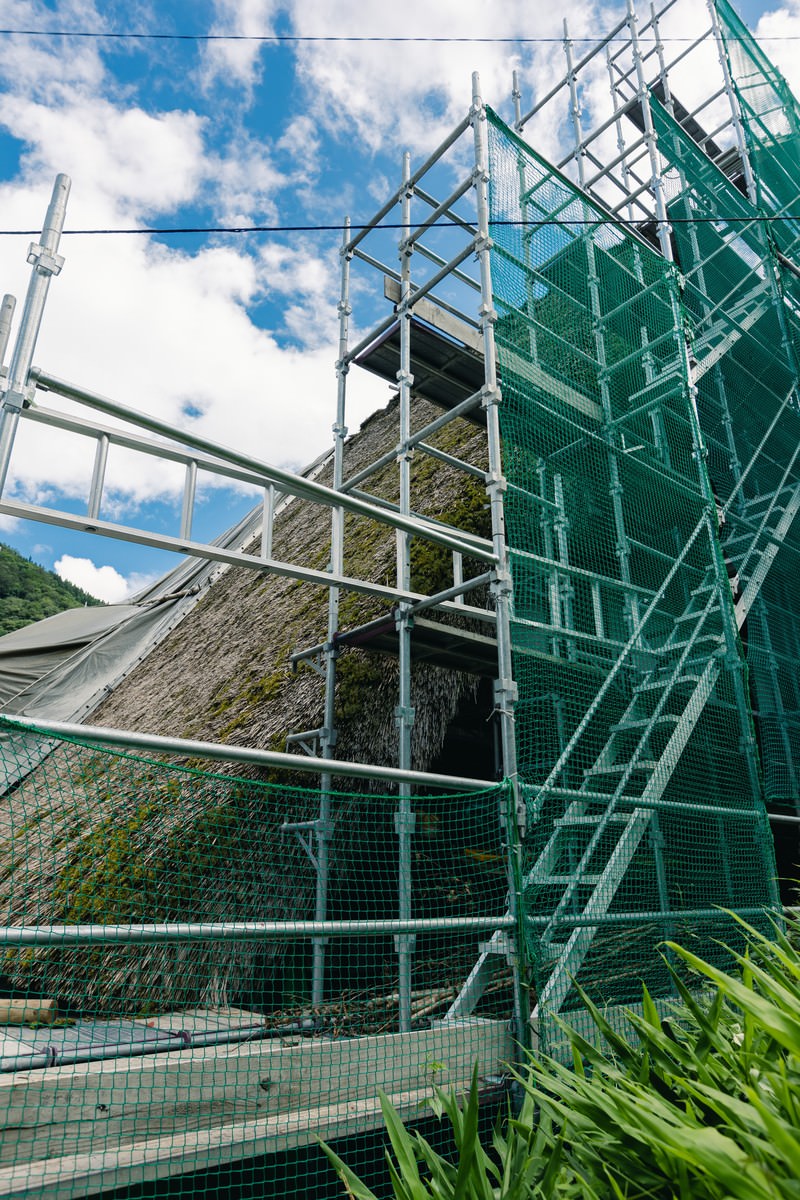 青空の下、緑色の足場が組まれた合掌造りの茅葺屋根建物
