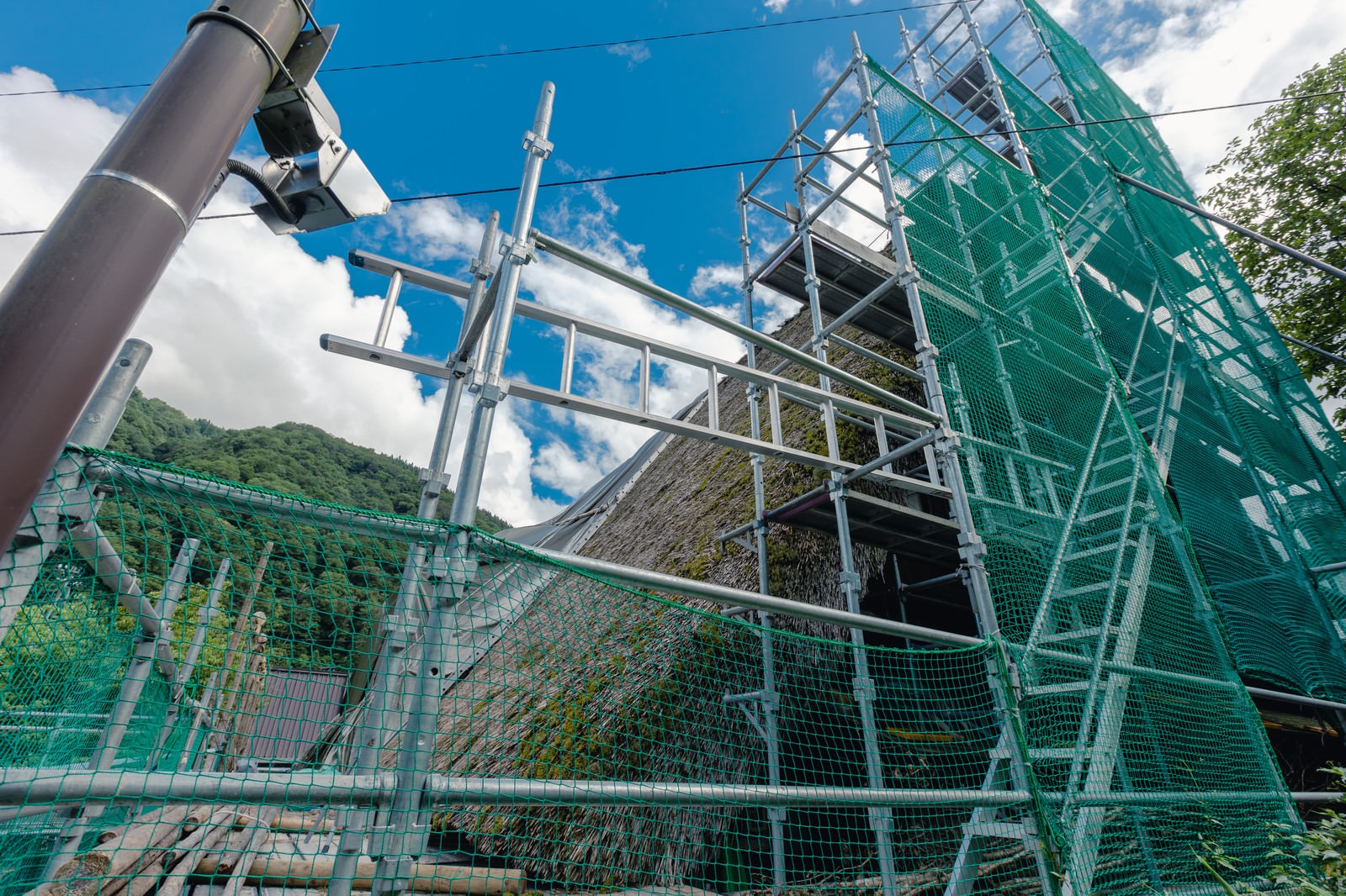 合掌造りの茅葺屋根に緑色の防護ネットで覆われた足場が組まれている