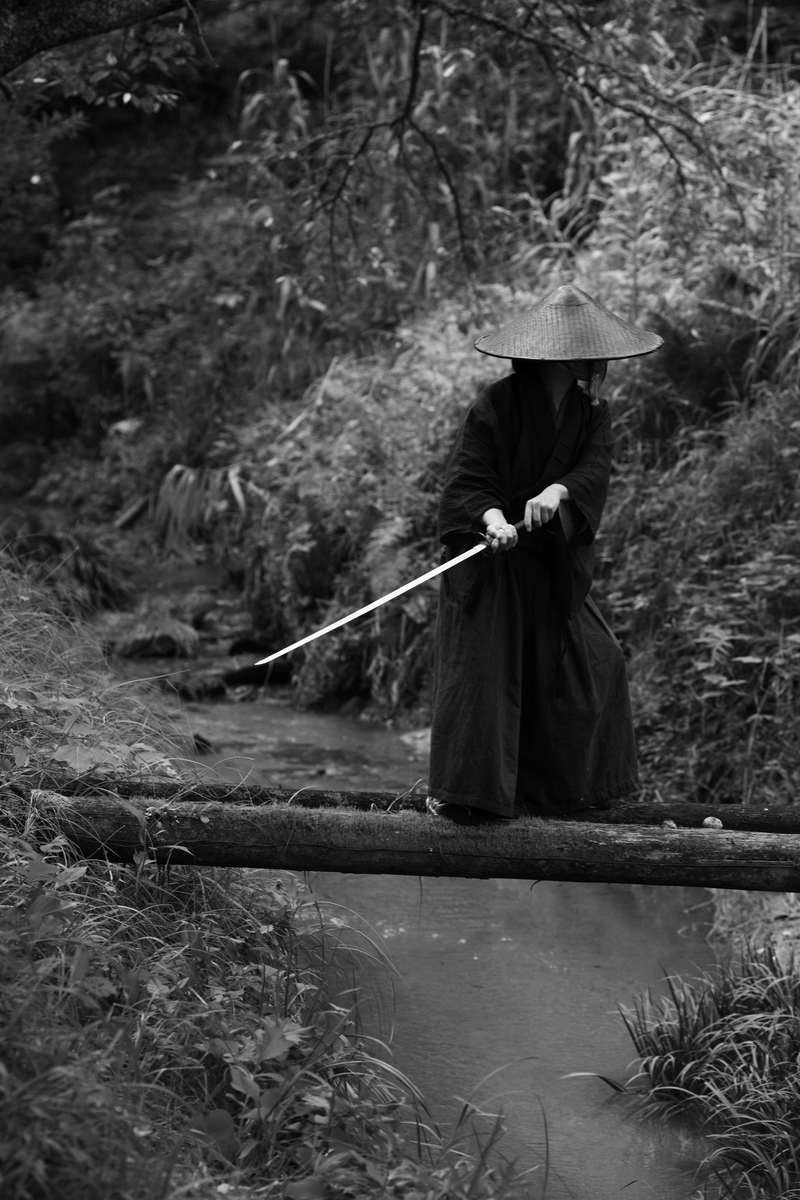 一本橋の上で刀を抜いて構える笠をかぶった袴姿の侍［モデル：まーこ］