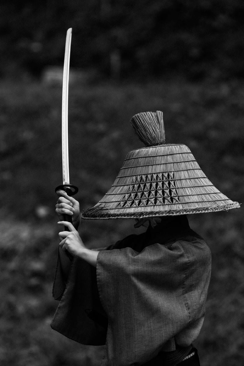 編み笠を被り袴を着た侍が日本刀を高く振り上げて上段の構えを取っている［モデル：まーこ］