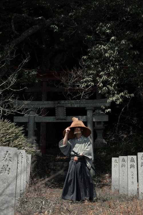 笠から零れ落ちる光、鳥居と奉納石柱前に立つ侍の武士