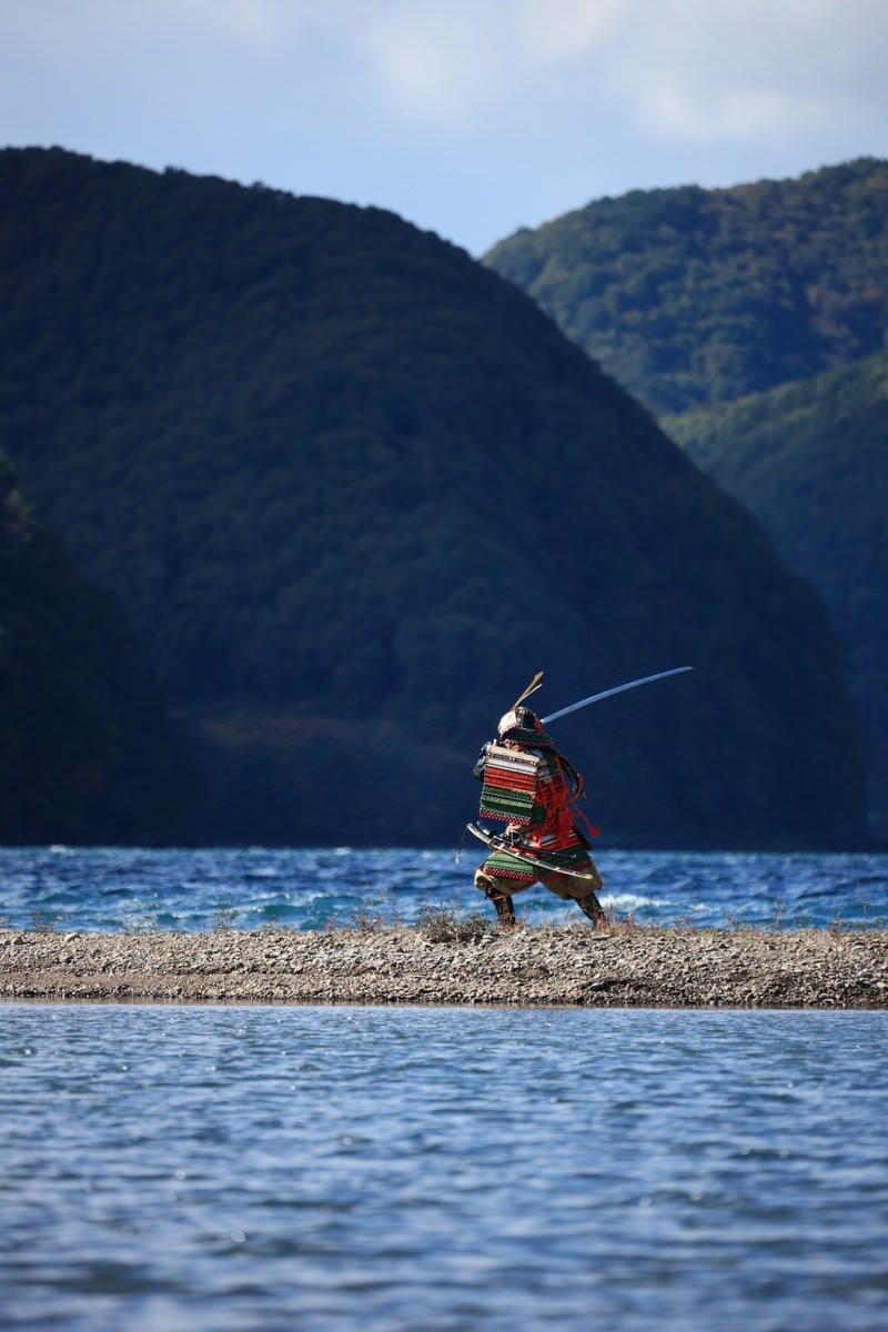 湖畔の中州で赤い大鎧を着て太刀を構える武士の後ろ姿［モデル：まーこ］