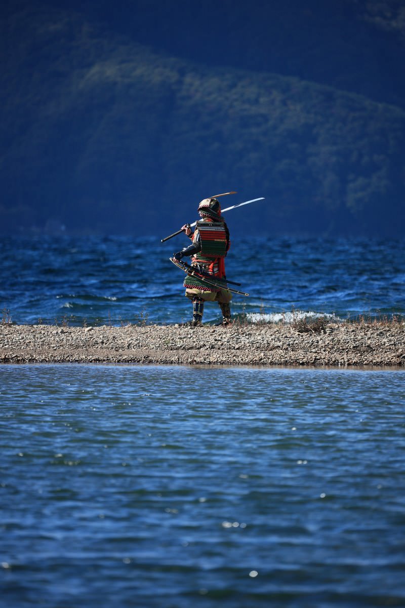 青い水辺の中州で赤い大鎧を着て日本刀を肩にかける侍［モデル：まーこ］