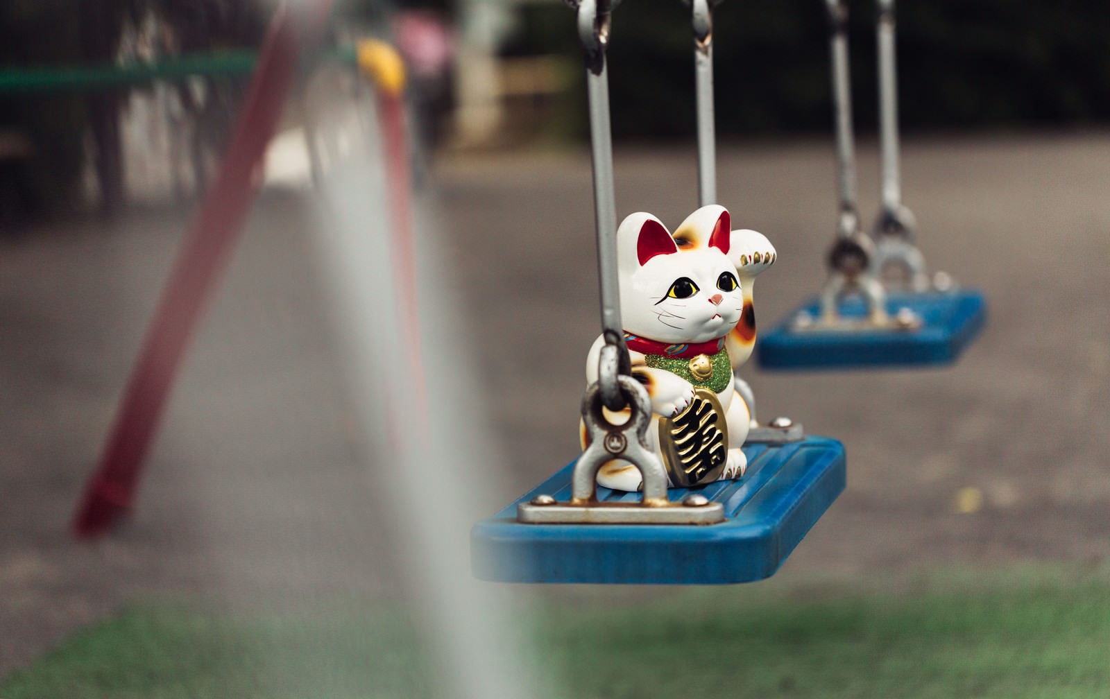 青いブランコの座板に乗った白い招き猫オブジェ。赤い耳と金色の腕、奥に複数のブランコが見える公園風景