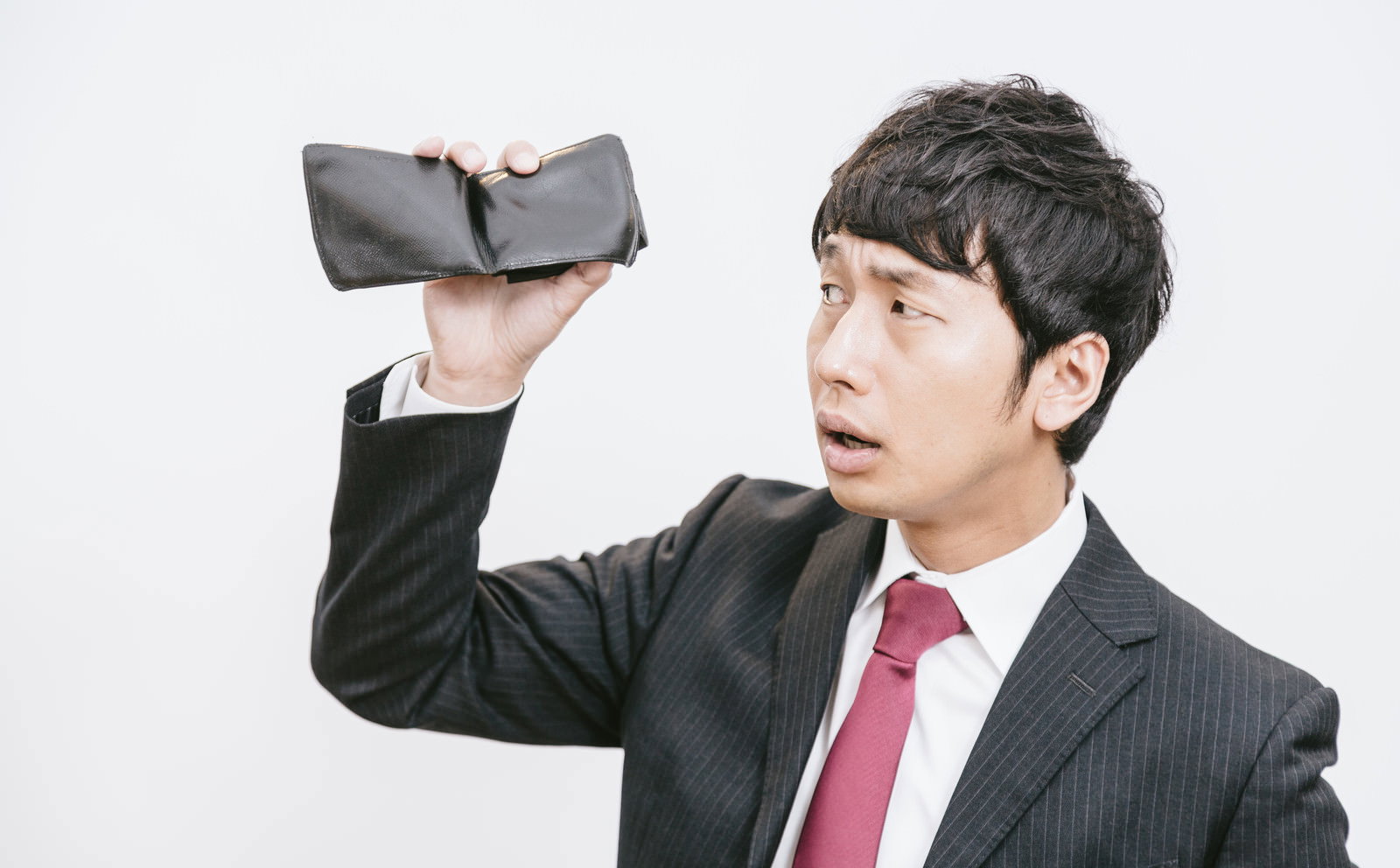 紺色のストライプスーツと赤紫のネクタイを着た男性が、開いた黒い財布を片手で持ち上げ、中身がないことに驚いた表情をしている［モデル：大川竜弥］