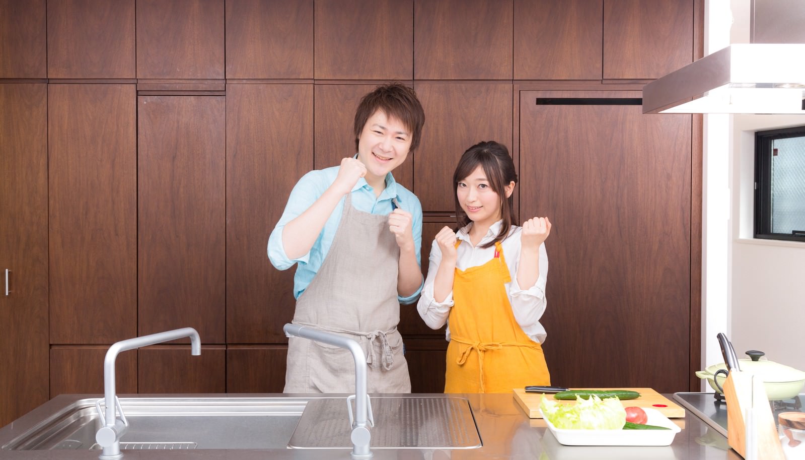 キッチンでエプロン姿の男女が笑顔でポーズをとる写真［モデル：五十嵐夫妻］