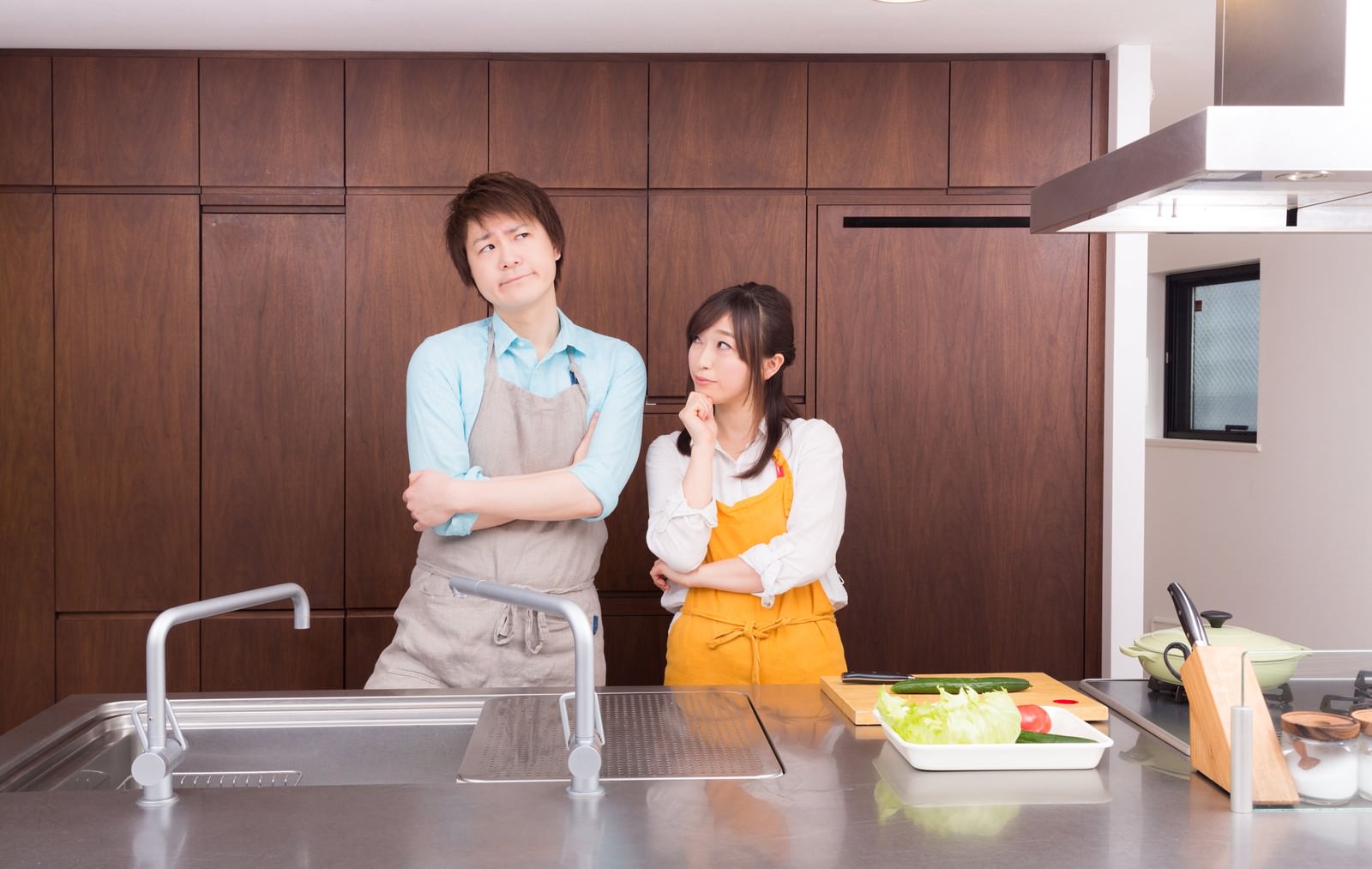 キッチンカウンターで夕食を考える黄色いエプロン姿の女性とベージュエプロンの男性［モデル：五十嵐夫妻］
