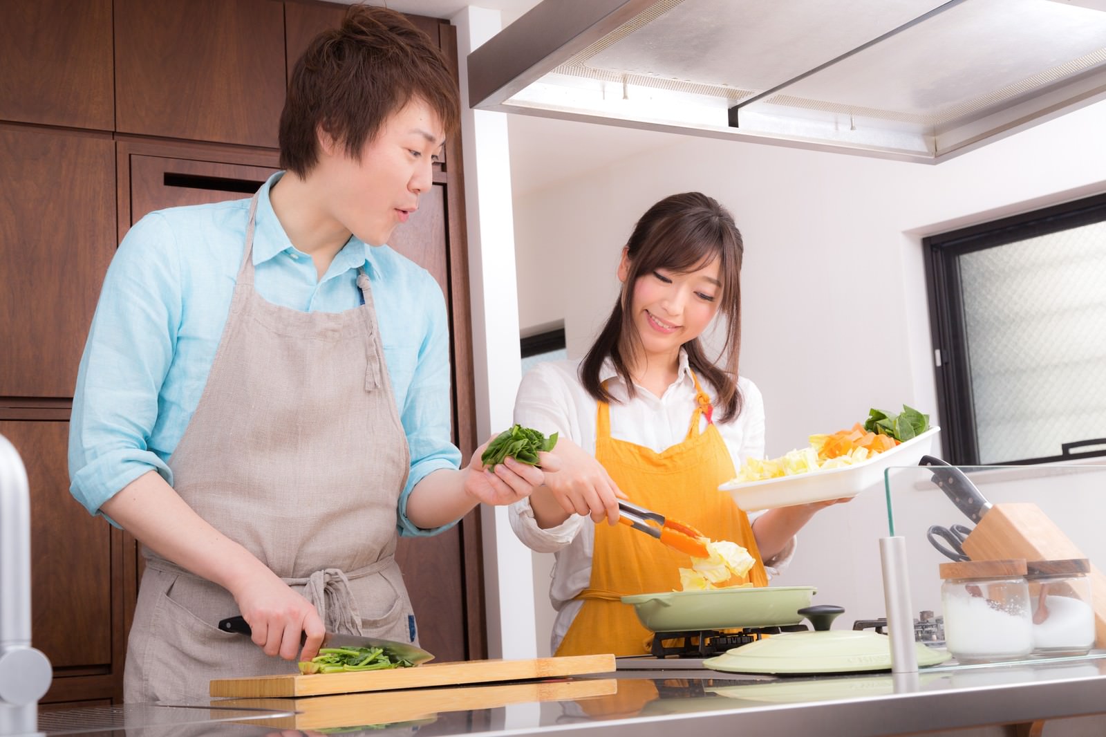 キッチンで野菜を切る男性と鍋の中身を確認する女性が笑顔で調理している［モデル：五十嵐夫妻］