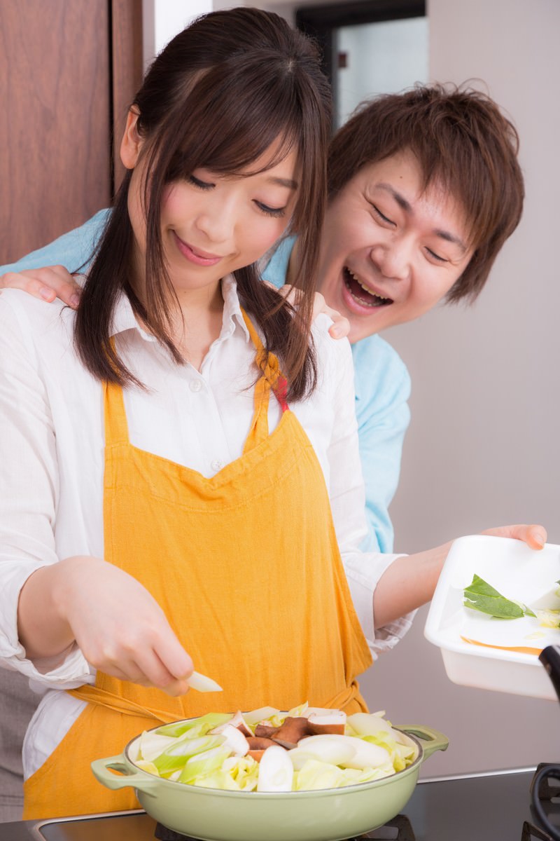 黄色いエプロンを着た女性が鍋に野菜を盛り付け、背後から男性が肩に手をかけて寄り添い笑う［モデル：五十嵐夫妻］