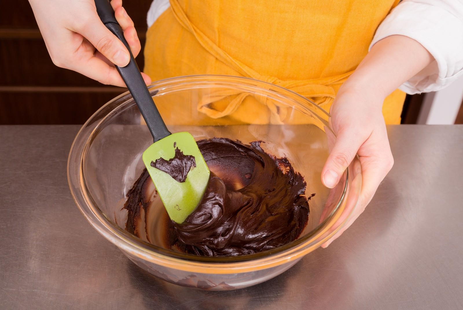 黄色いエプロンの人物がボウルのチョコレートをゴムベラで練っている様子［モデル：五十嵐夫妻］