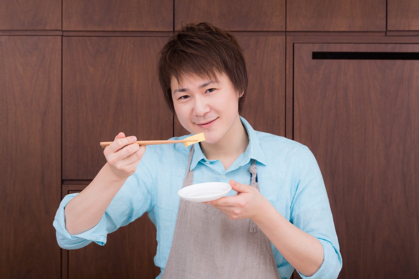 玉子焼きを箸で持ち、評価する表情を見せる男性。白い皿と木目調の背景［モデル：五十嵐夫妻］