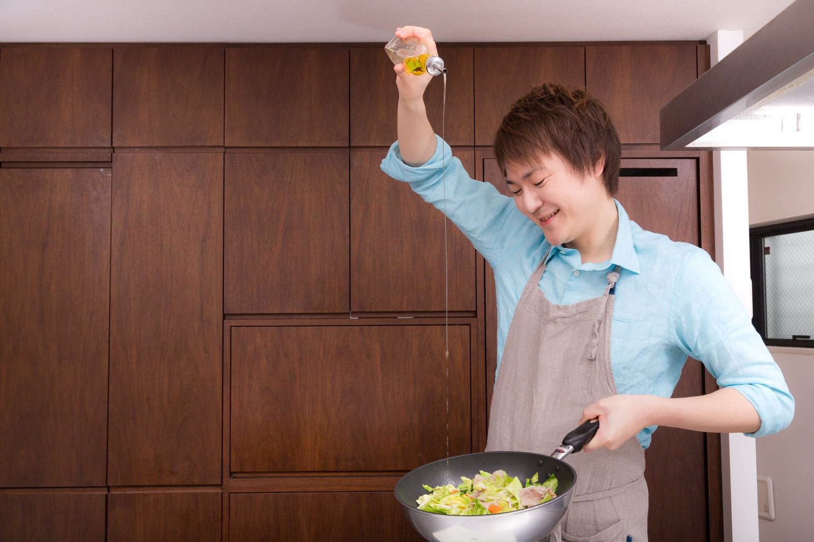 キッチンの前で野菜炒めにオリーブオイルを注ぐ笑顔の男性［モデル：五十嵐夫妻］