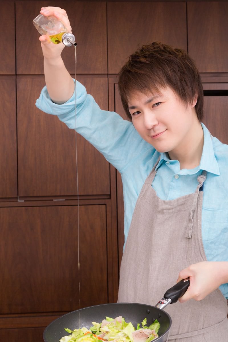 エプロン姿の男性がフライパンの野菜炒めに透明な瓶からオリーブオイルを注いでいる調理風景［モデル：五十嵐夫妻］