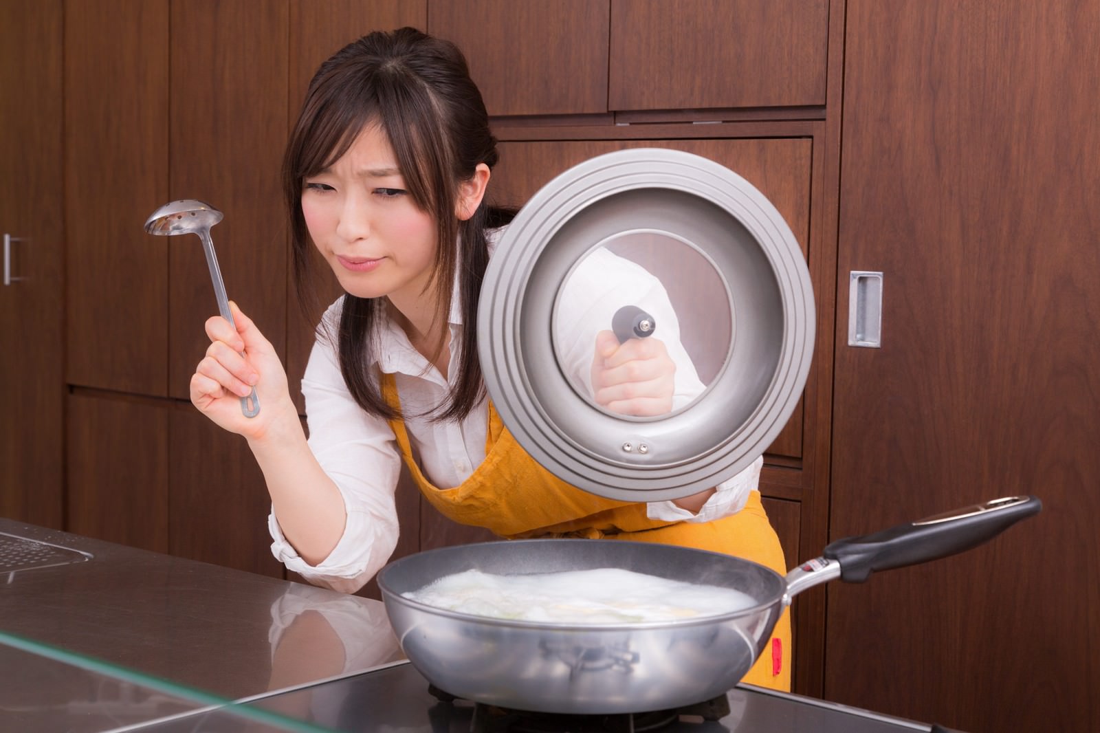 黄色いエプロン姿で鍋の蓋とフライパンを手に調理を確認する女性とキッチン［モデル：五十嵐夫妻］