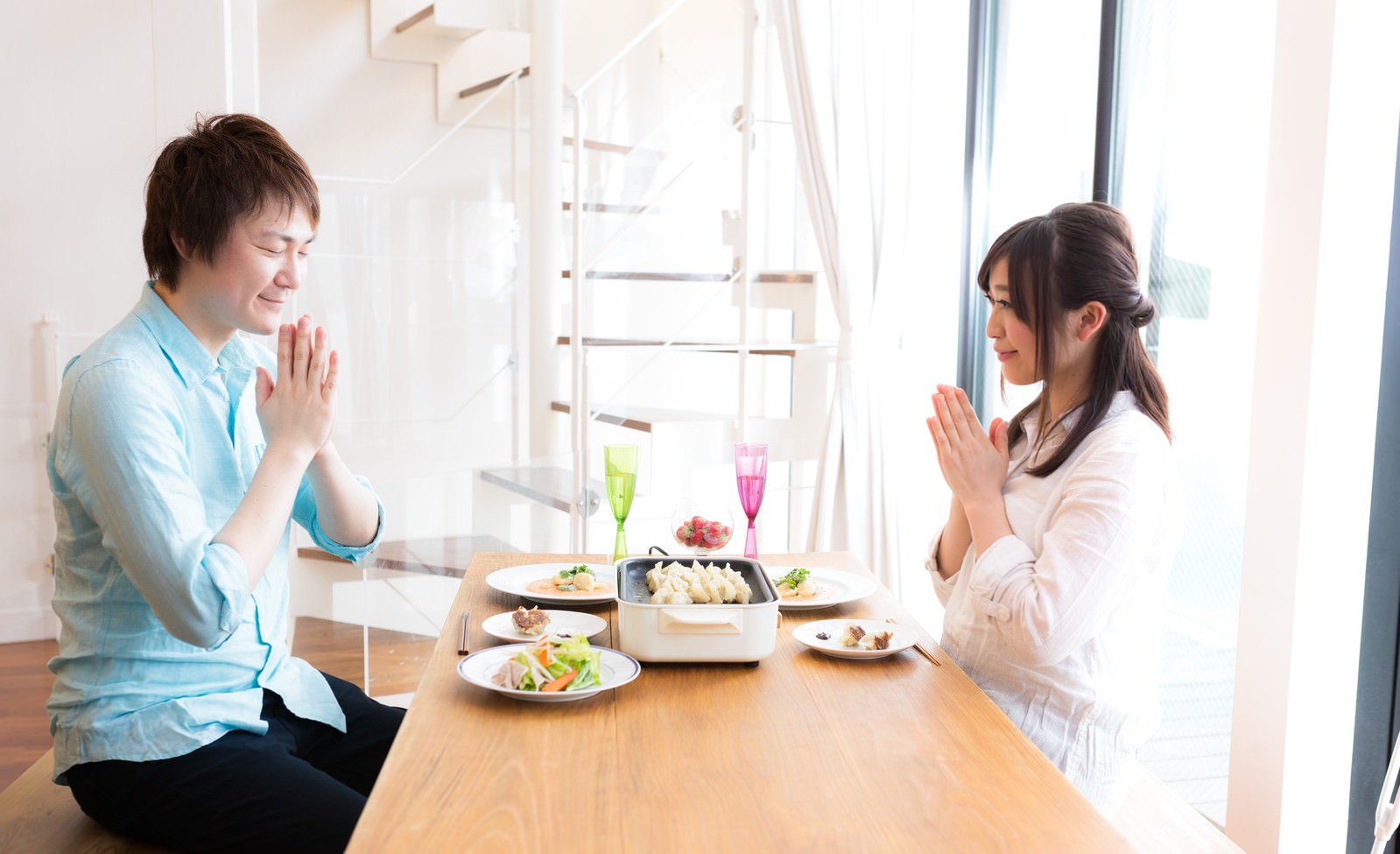 木製テーブルで両手を合わせて食事を始める夫婦の写真［モデル：五十嵐夫妻］