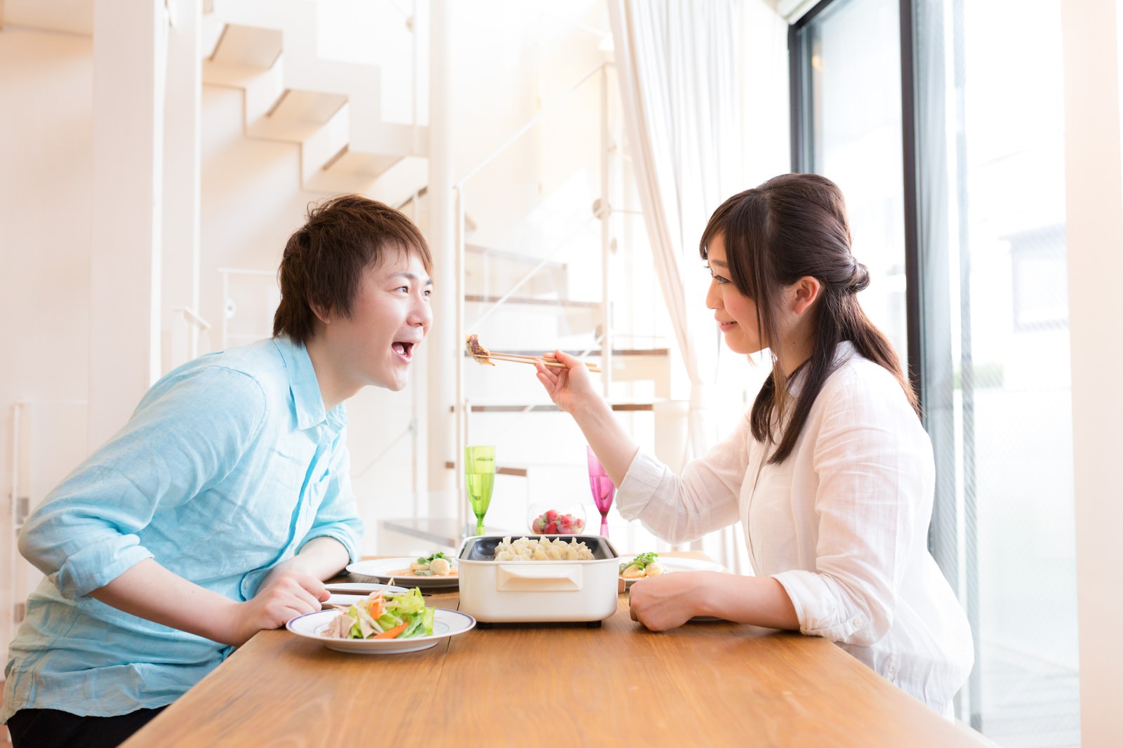 窓際で食卓に向かい、女性が男性に箸で餃子を食べさせている夫婦の様子［モデル：五十嵐夫妻］