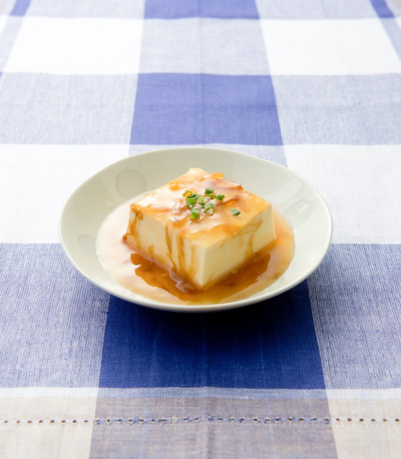 白い丸皿に四角い豆腐にオレンジ色のあんかけと刻みネギがかかっている