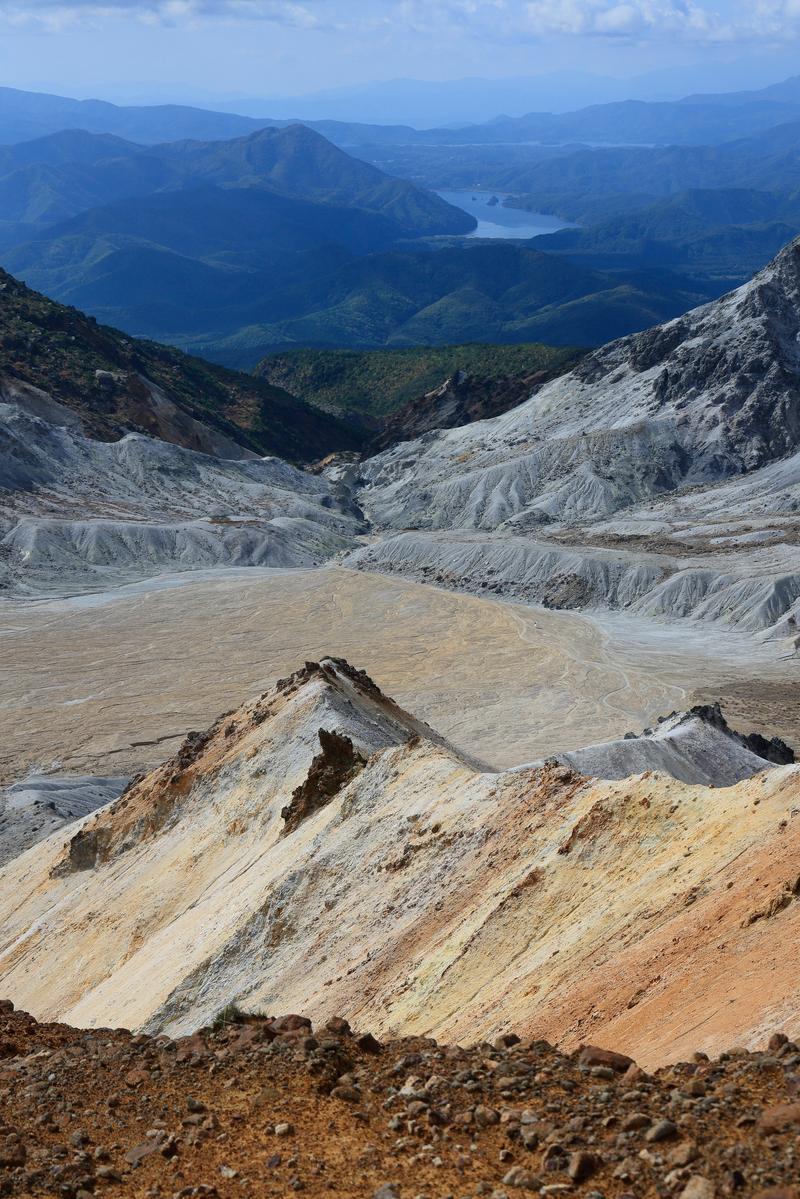 沼ノ平火口の褐色の火口岩肌が露出した荒涼とした地形。背景に緑の山々と青い湖がある