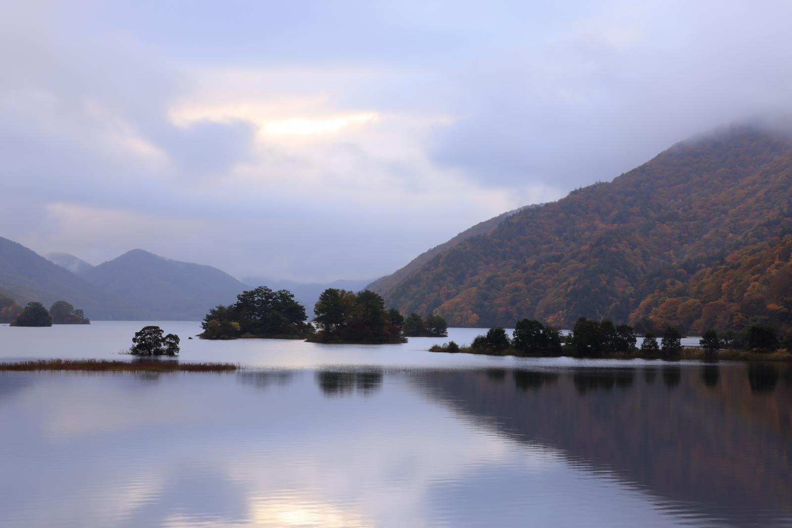 秋元湖の湖面に映る紅葉の山々。朝焼けの空と静寂な水面が印象的。