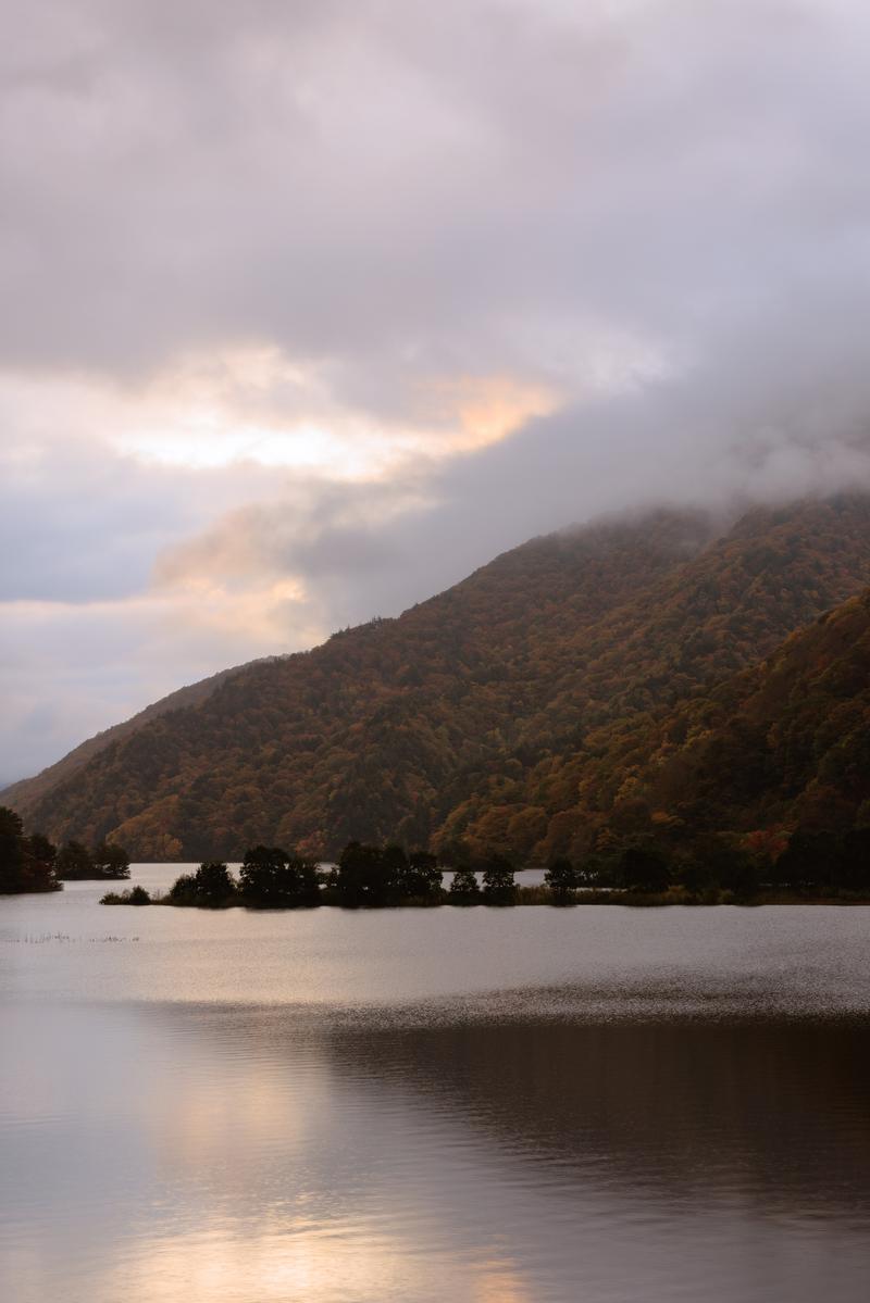 雲間から朝焼けが見える秋元湖の風景。紅葉に染まった山が湖面に映り込んでいる
