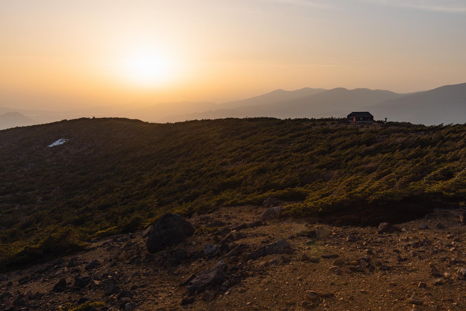 夕焼けに染まる山々を背景に、稜線に建つ鉄山避難小屋