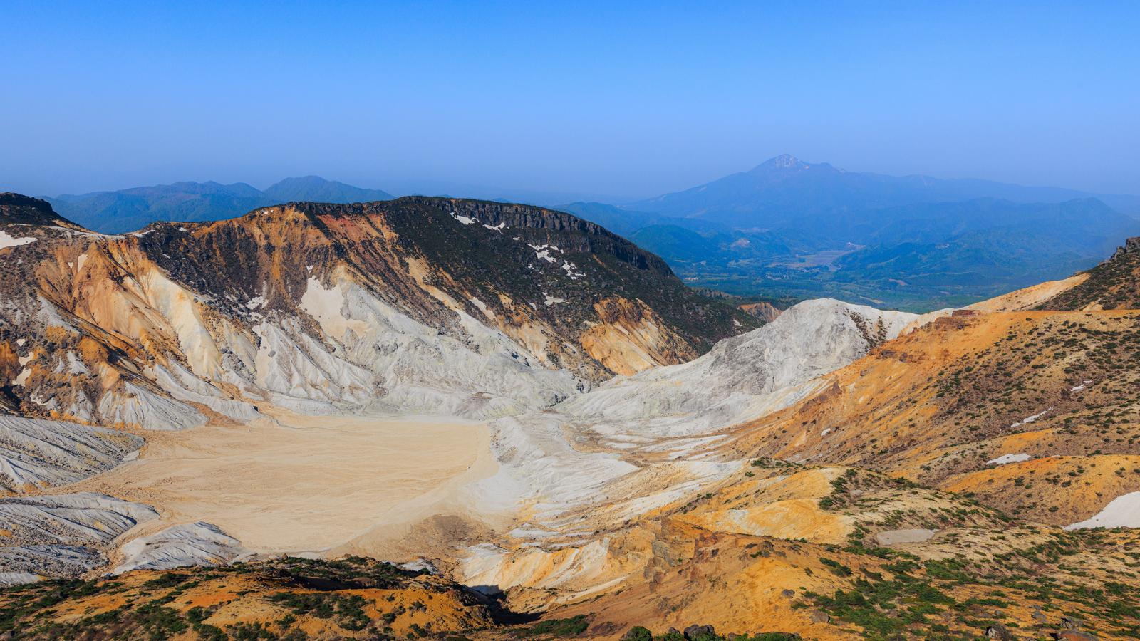 白い岩肌が露出した火口を手前の新緑が彩る火山景観