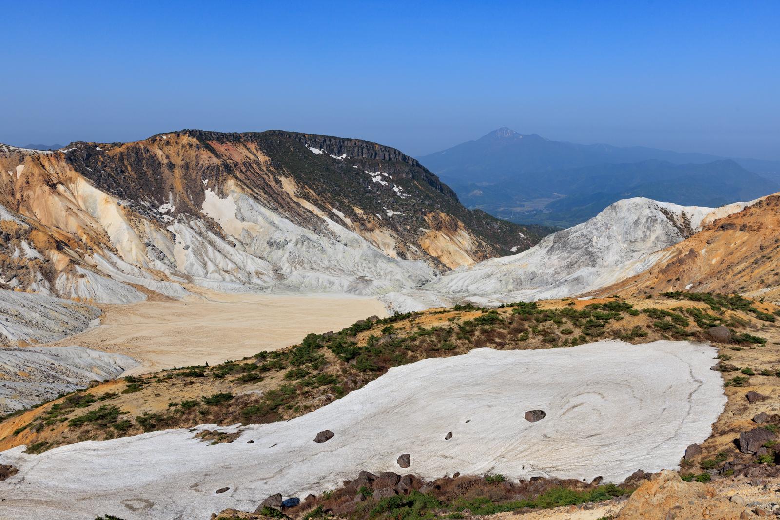 白い硫黄地と焦げ茶色の火山岩からなる沼ノ平火口。手前に新緑の低木が生育。