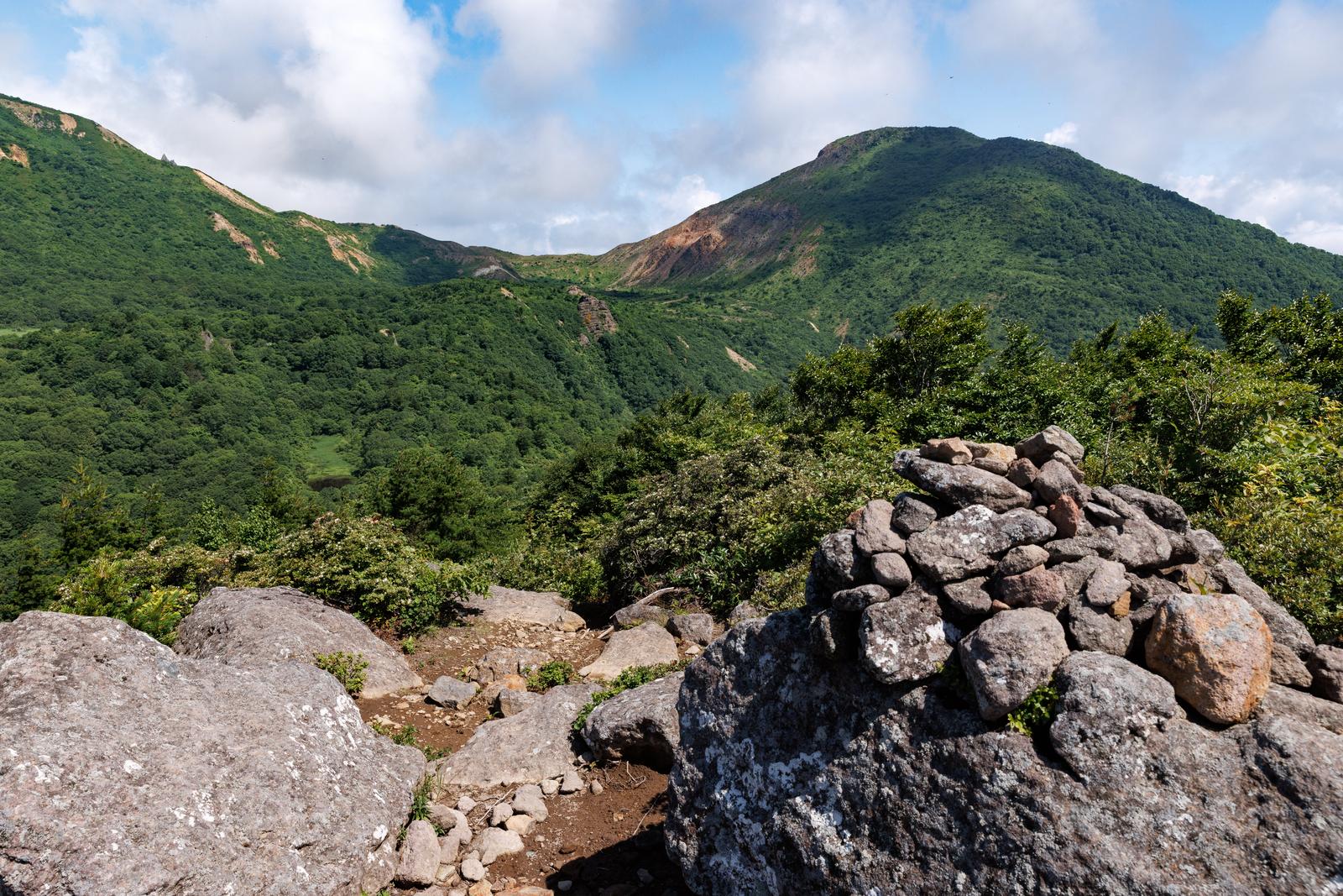 山頂のケルンと遠くに連なる磐梯山の山々、緑の谷間を見下ろす風景