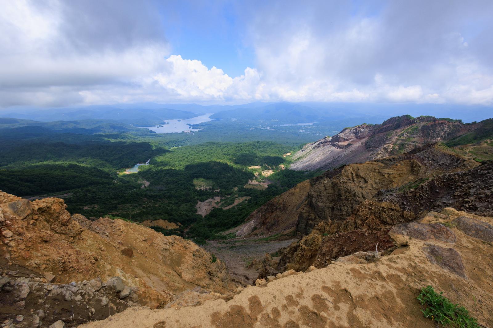 磐梯山登山口の露出した火山岩と緑の森、遠方の湖が見える山岳風景