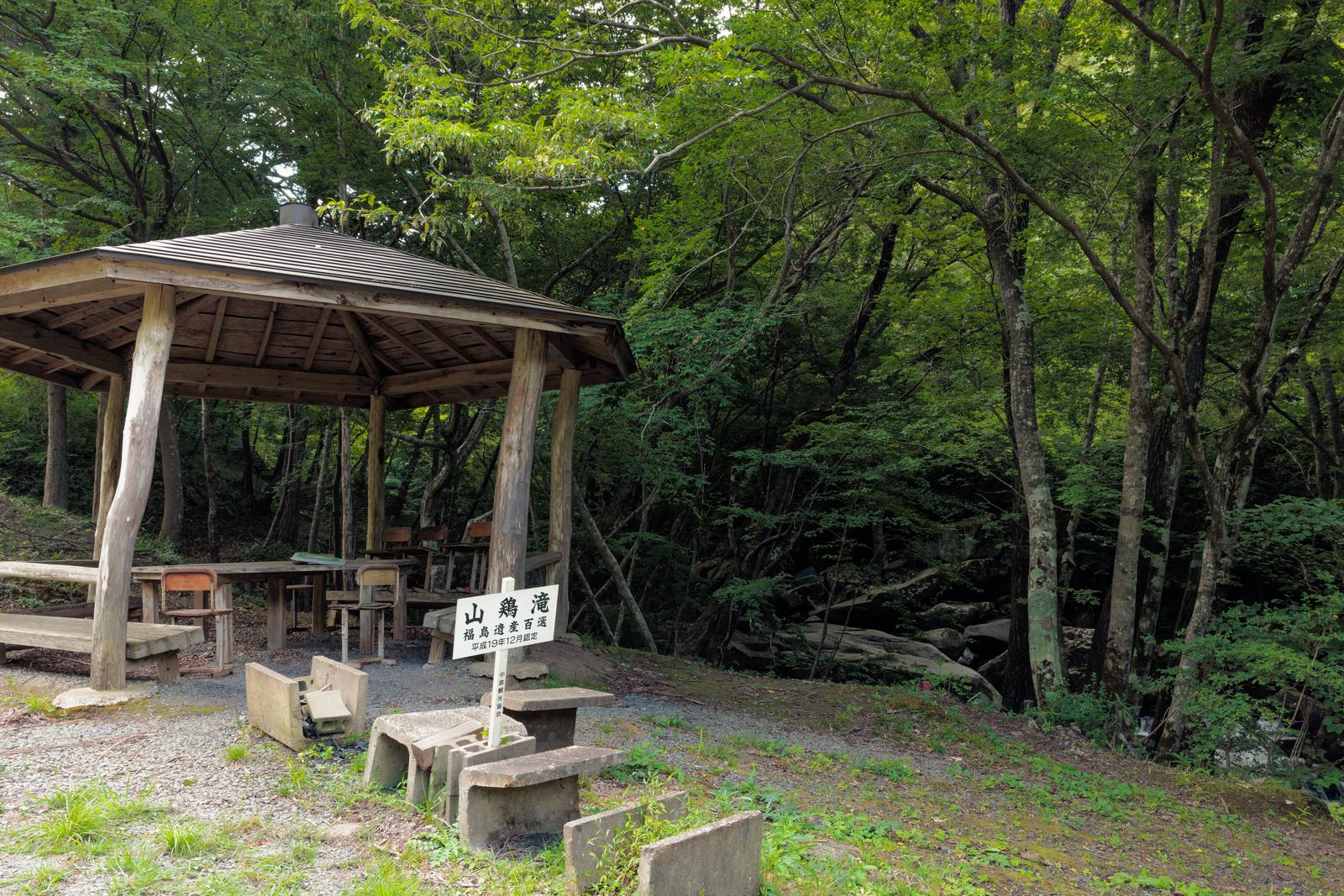 山鶏滝の森の中に建つ木造の東屋と白い標識、石製ベンチ