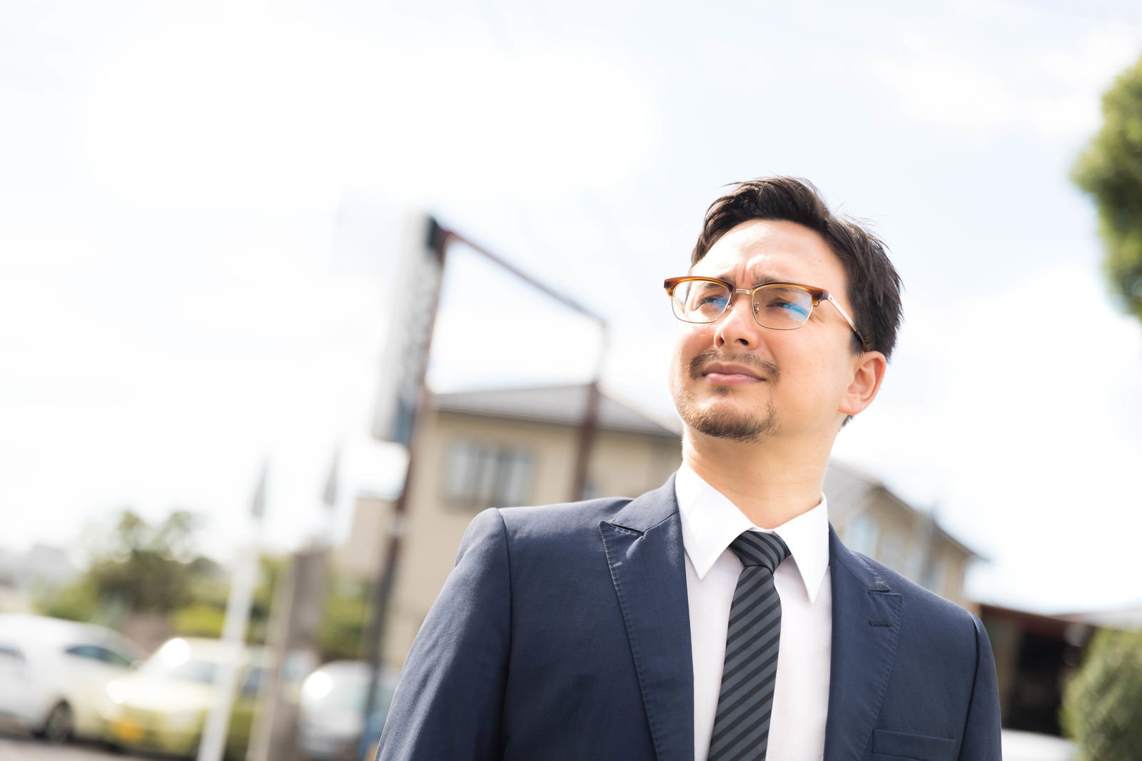 紺色のスーツとネクタイを着けた眼鏡の男性が屋外で上を向いて立っている［モデル：Max_Ezaki］
