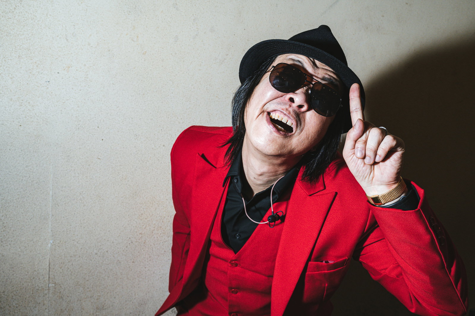 赤いスーツと黒い帽子、サングラス姿で指を立てて話しかける男性［モデル：のせさん］
