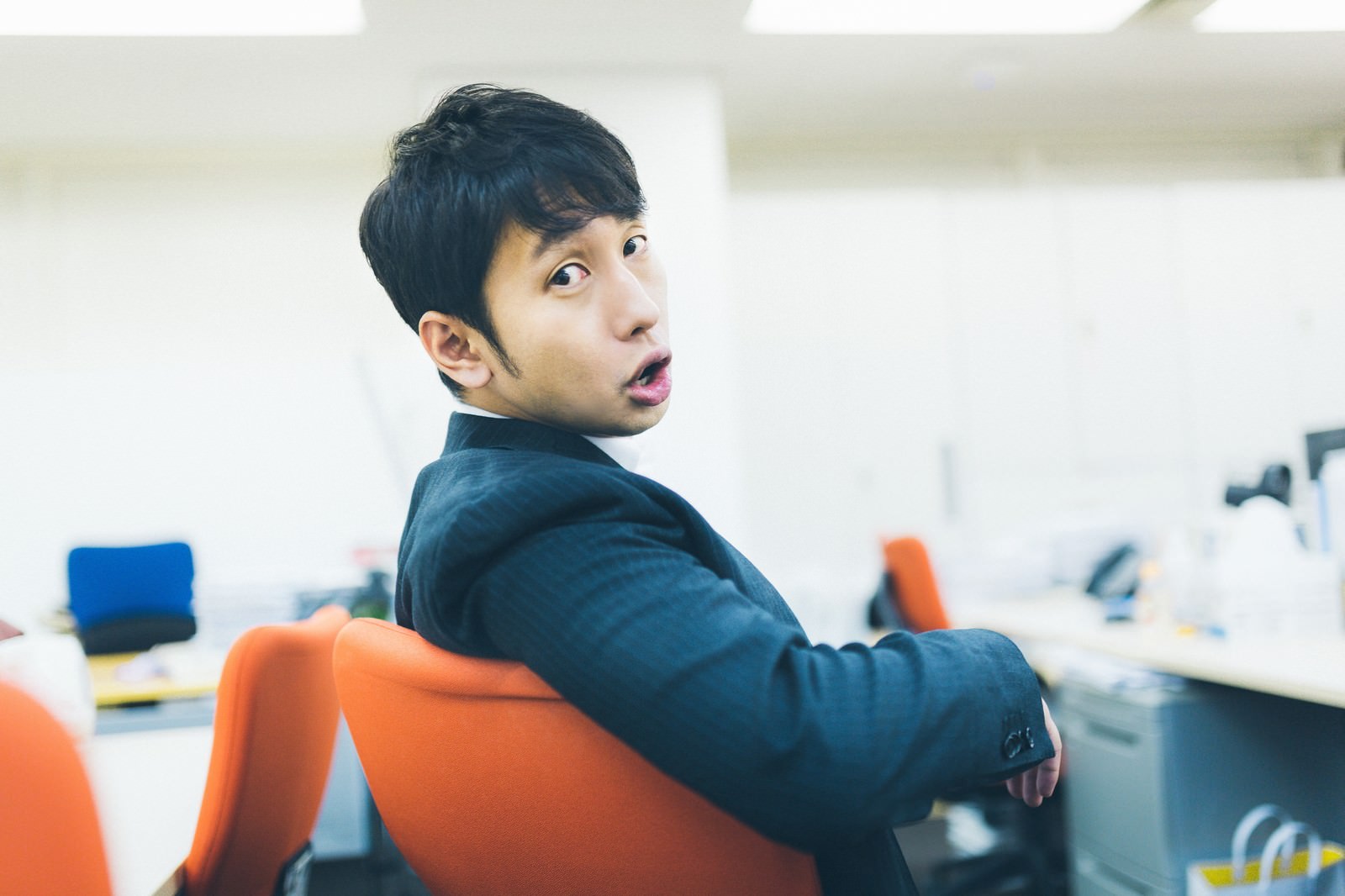 オフィスのオレンジチェアで肩越しに振り向き、指導する紺スーツ姿の男性上司［モデル：大川竜弥］