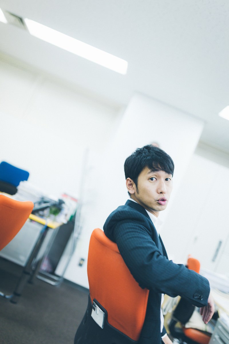 オレンジチェアに座るスーツ姿の男性がカメラを向きながらアヒル口をしている［モデル：大川竜弥］