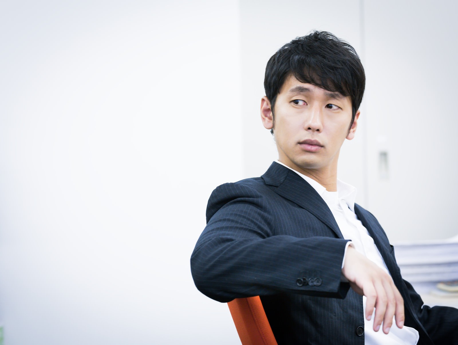 紺色ストライプスーツを着た男性がオレンジの椅子に座り、身体を後ろに向けている［モデル：大川竜弥］