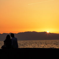夕暮れの水辺に並んで座る二人のシルエットの写真
