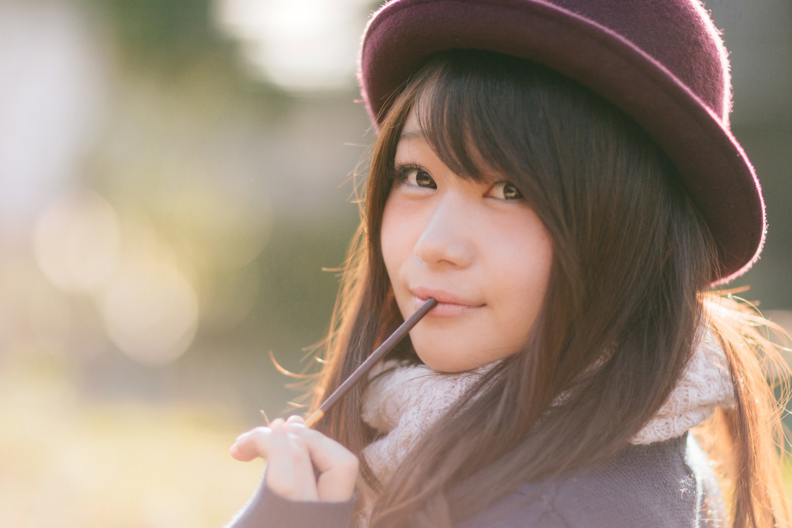 ピンク色のベレー帽を被った女性がポッキーを口にくわえ、暖色の逆光の中で上を向いている［モデル：茜さや］