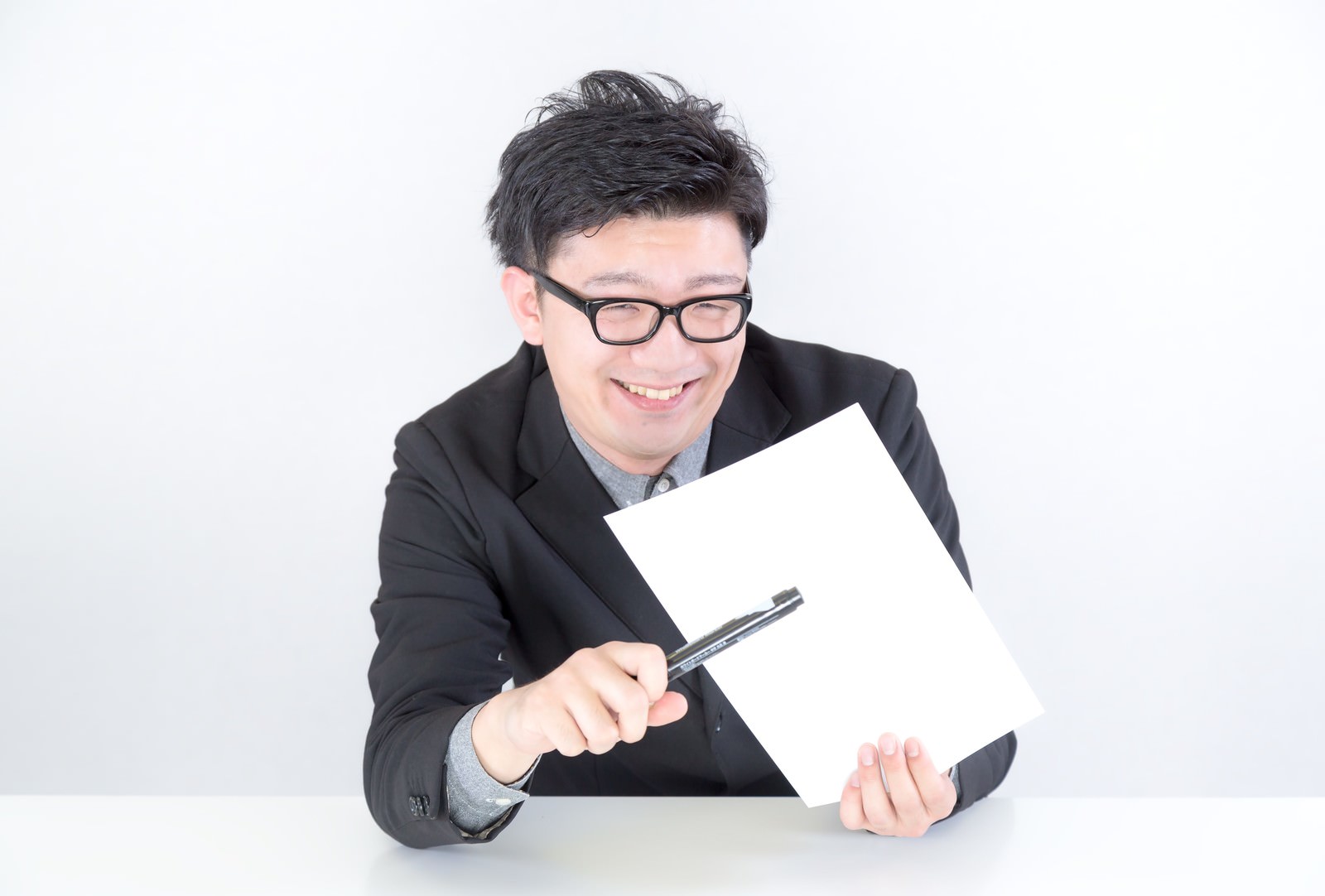 黒いスーツと眼鏡をかけた男性が白い机で書類を見下ろし笑顔で握り持つ［モデル：OZPA］