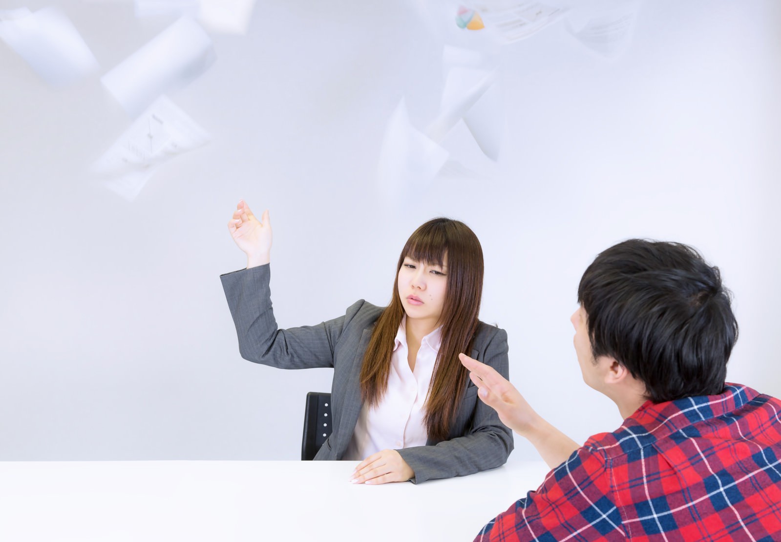 テーブル越しにプレゼンする女性デザイナーと指摘する男性、散乱する資料［モデル：大川竜弥 Lala］