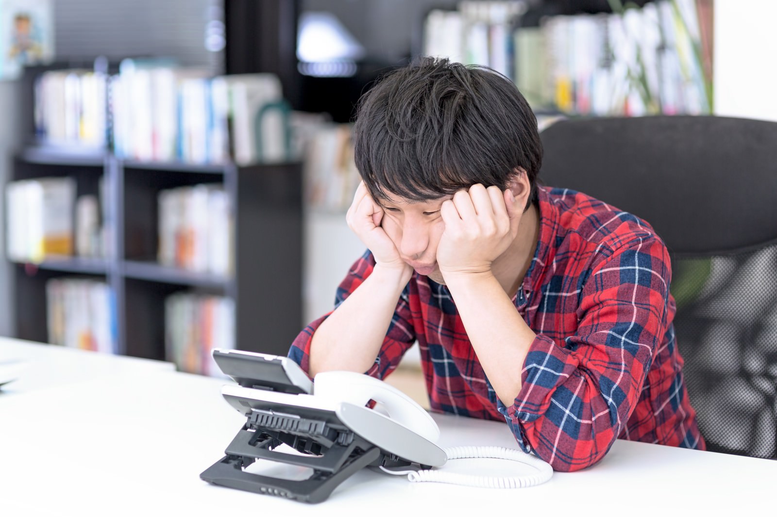机に向かい両手で顔を抱える赤チェック柄シャツの男性、手前にスマートフォン、背後に本棚［モデル：大川竜弥］