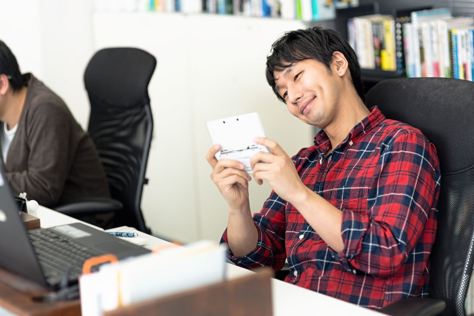 オフィスのデスクでゲームパッドを握り画面を見つめるチェック柄シャツの男性［モデル：大川竜弥］