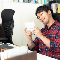 携帯ゲーム機を持って本棚の前に座る男性の写真